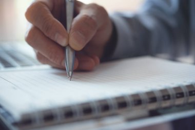 Business man hand write his work plan on paper notebook with a pen while working in modern office. Man student taking note while learning online course via laptop computer, close up, elearning concept