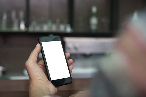 Man hand holding blank white screen mobile smartphone in coffee shop with blurred background of wooden counter bar in brown coffee tone, close up
