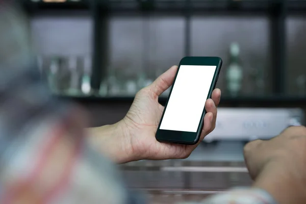 Man hand holding blank white screen mobile smartphone in coffee shop with blurred background of wooden counter bar in brown coffee tone, close up