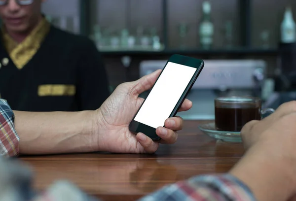 Mock up image, man hand holding blank white screen mobile smartphone in coffee shop with blurred background of wooden counter bar and male barista in brown coffee tone, close up, vintage tone