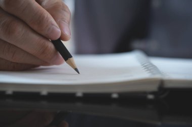 Business man, project manager with black pencil in hand writing on paper notebook, project planning on desk in office, close up, front view