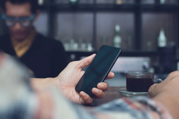Man sitting at counter bar and holding blank copy space screen mobile smartphone with blurred male barista in coffee shop, close up