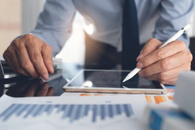 Business strategy analysis concept. Business man with digital pen analyzing marketing report, pointing at financial graph on digital tablet and working on laptop computer with spreadsheet on desk in modern office, close up