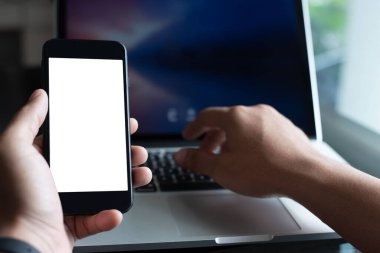 Mock up, man hand using blank white screen mobile amart phone, working on laptop computer on office desk, use for internet payment, mobile application or web design template