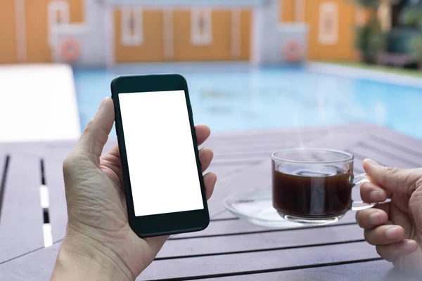 Mock up image of casual man using blank white screen mobile smartphone and drinking black coffee near swimming pool at modern luxury hotel. Man checking internet application on phone with copy space, modern lifestyle concept
