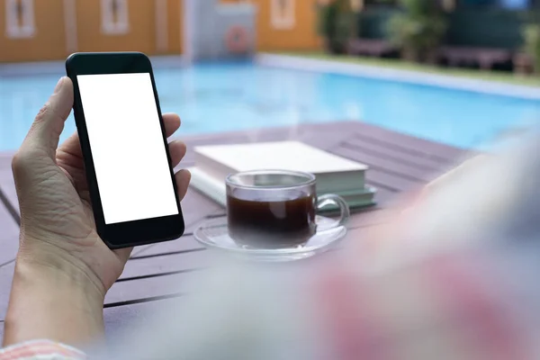 Mock up image of casual man using blank white screen mobile smartphone and drinking black coffee near swimming pool at modern luxury hotel. Man checking internet application on phone with copy space, modern lifestyle concept