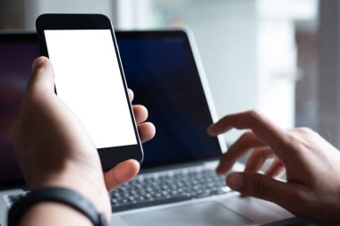Mock up, man hand using blank screen smart mobile phone, working on laptop computer on office desk as background for internet payment mobile apps or web design, product advertising