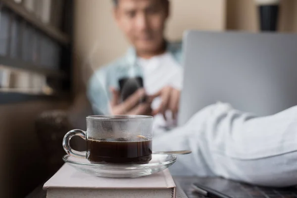 Hot coffee cup with steam on desk with blurred casual businessman or hipster using mobile smartphone and laptop computer at home