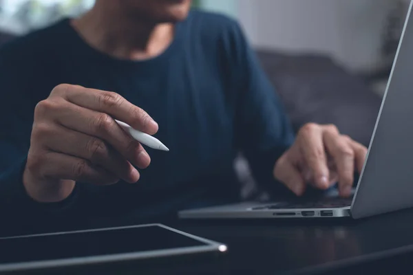Website designer with stylus pen working digital tablet and computer laptop on wooden desk. Casual business man hand touching on touchpad screen, close up. Freelance online working from home, close up