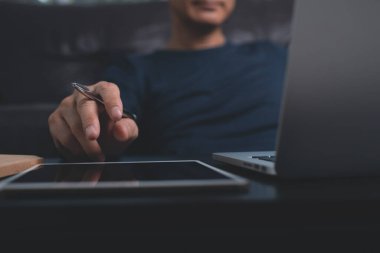 casual business man with pen in hand sitting at wooden desk, looking on laptop computer screen with digital tablet on wooden desk. Freelancer online working from home, close up. Business and technology concept