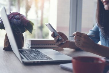 Başarılı Asyalı iş kadını cep telefonuyla kalem tarama interneti kuruyor, bilgisayarla çalışıyor, ofis masasında bir fincan kahve ile ofis masasında çalışıyor,
