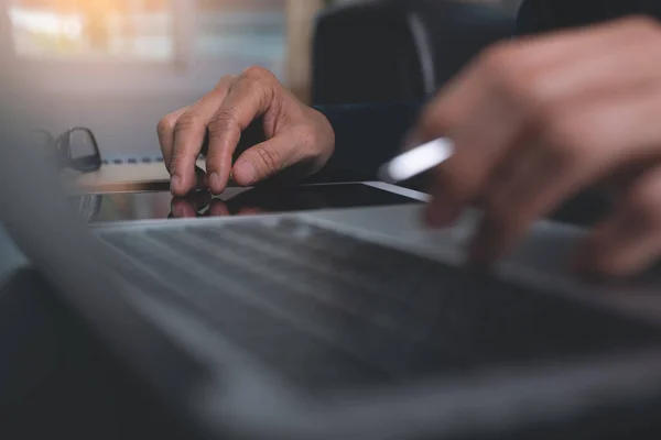 Business man with pen in hand using and touching on digital tablet screen while working on laptop computer on wooden desk in modern office, close up, dark tone. Online working, E-business technology concept.