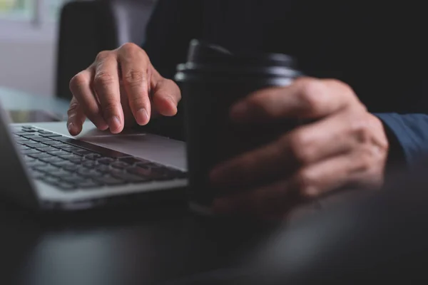 Business man hand working on laptop computer with cup of coffee on wooden office desk. Freelance networking, browsing internet at home, close up, dark tone. Online working concept