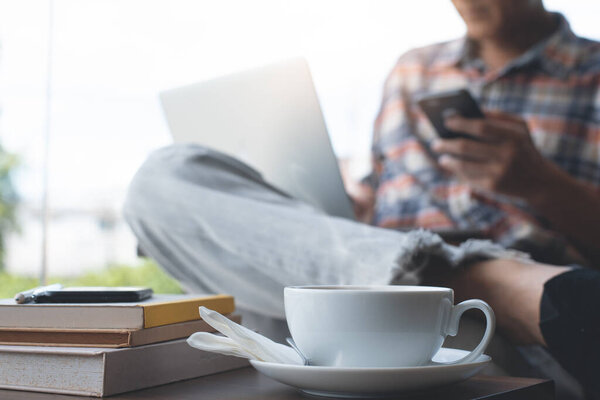 Casual man, hipster, freelance using mobile smart phone, working on laptop computer with cup of coffee and books on wodden table at coffee shop, close up. Online working, portable office concept