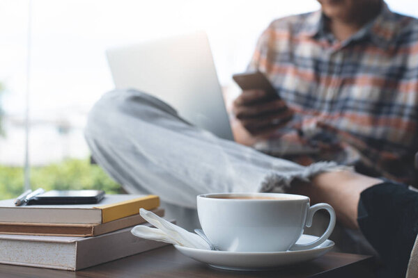 Casual man, hipster, freelance using mobile smart phone, working on laptop computer with cup of coffee and books on wodden table at coffee shop, close up. Online working, portable office concept