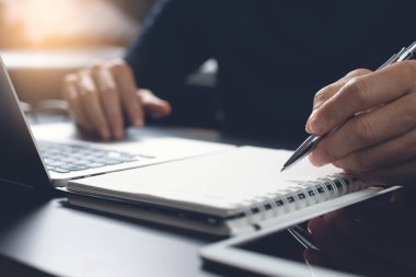 E-learning, online education. Student writing content on paper notebook with a pen while studying online course via laptop computer with digital tablet on desk. Business man planning work on notepad in office. Startup business, close up