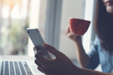 Cropped, business woman, entrepreneur holding red cup of hot coffee, morning news update via smart phone application, working on laptop computer in office, social media communication concept