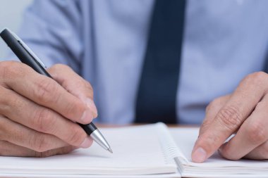 Businessman sitting at office desk writing something on paper notebook, close up.