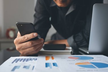Buisness accounting. Man accountant using mobile phone and working on laptop computer. Businessman reviewing business marketing financial report on desk in modern office, copy space, toned, close up