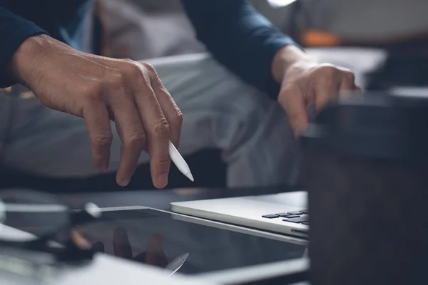 Casual business man with stylus pen writing on digital notepad, touching on digital tablet screen, working on laptop computer in office. Graphic or web designer working his project, dark tone, close up. Startup business, technology concept