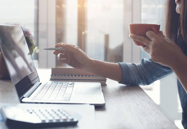 Cropped, smart asian businesswoman relaxed working on laptop computer and drinking coffee in modern office near the window with calculator and business report on office desk