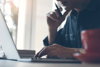 Casual business man or freelancer working on laptop computer, thinking about his project planning, holding a pen with cup of coffee on desk in home office, close up