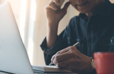 Casual business man, freelancer calling via mobile phone, working on laptop computer smart phone with cup of coffee on desk, online working from home office, close up.