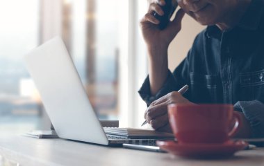 Casual business man, freelancer calling via mobile phone, working on laptop computer smart phone with cup of coffee on desk, online working from home office, close up.