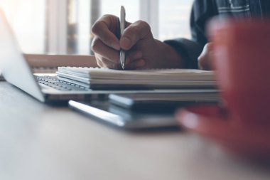 Casual business man writing on notepad, working on laptop computer with smart phone, cup of coffee on wooden table in the morning at home office, close up