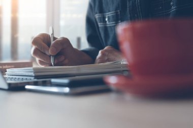 Casual business man writing on notepad, working on laptop computer with smart phone, cup of coffee on wooden table in the morning at home office, close up