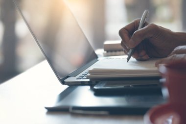Man, freelancer writing on notepad, working on laptop computer with smart phone, digital tablet, cup of coffee on table at home office or coffee shop, close up