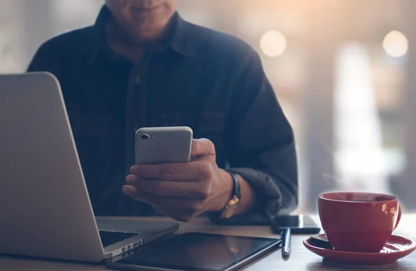 Businessman, freelancer using mobile smartphone, working on laptop computer. Man browsing internet via smart phone with cup of coffee on table, online working from coffee shop, close up.