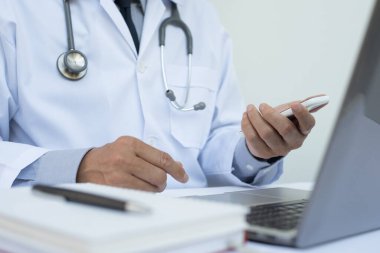 Cep telefonu kullanan erkek doktorların kapatılması ve hastanedeki beyaz masadaki dizüstü bilgisayarda telif alanı, elektronik sağlık kayıt sistemi EHR, telekonferans veya teletıp konsepti ile çalışılması.