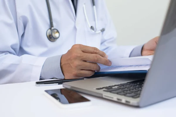 Close up of male doctor reading patient health record chart and working on laptop computer on white desk in hospital with copyspace, electronic health records system EHRs, teleconference or telemedicine concept.