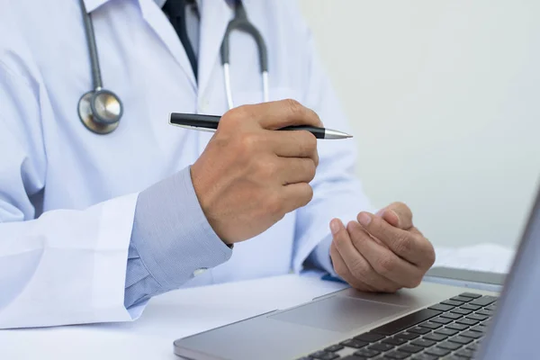 Erkek doktorun elinde kalemle hastanenin beyaz masasındaki dizüstü bilgisayarda fotokopi odası, elektronik sağlık kayıt sistemi EHR, telekonferans veya teletıp konsepti ile çalışmasını kapatın..