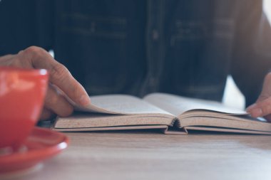 Sabahleyin pencerenin yanındaki tahta masada kırmızı kahve fincanı olan bir adam kitap okuyor.