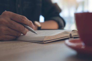 Ahşap masada elinde kalem ve bir fincan kahveyle kitap okuyan adam.