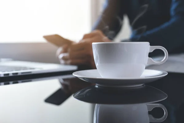 Cup of hot coffee and smoke on table with casual man using mobile smart phone and working on laptop computer as background with morning sunshine, copy space