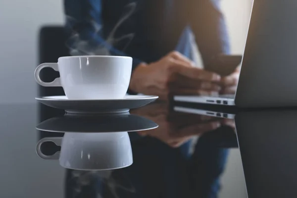 Cup of hot coffee and smoke on table with casual man using mobile smart phone and working on laptop computer as background with morning sunshine, copy space