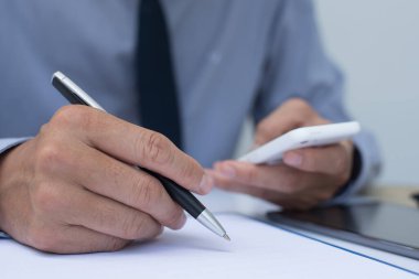 İş adamı tükenmez kalemle not defterine yazı yazıyor ve ofis odasında cep telefonu tutuyor. Çevrimiçi çalışma konseptini kapatıyor..