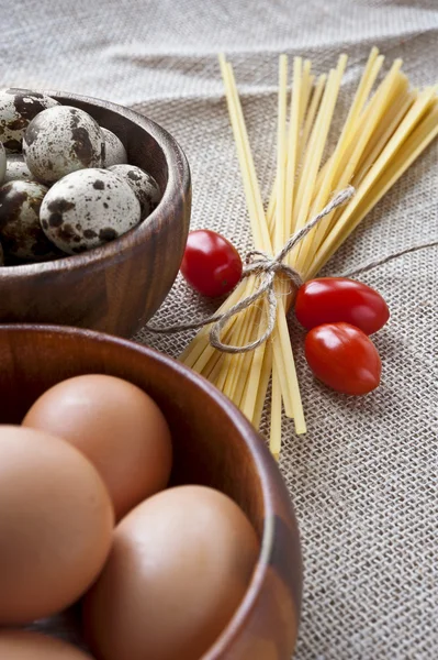 Dry pasta, tomatoes and eggs in wooden bowls on sack background — Stock Photo, Image