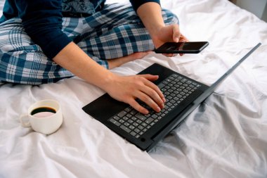 Genç adamın yatakta dizüstü bilgisayar kullanırken görüntüsünü kes. Üzerinde pijama giymiş bilgisayar klavyesi kullanan tanınmayan bir erkek öğrenci var.