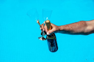 Mavi suya karşı elinde iki flüt bardağıyla şampanya şişesi tutan bir erkek. Havuz başında kutlama etkinliği. Lüks yaşam tarzı