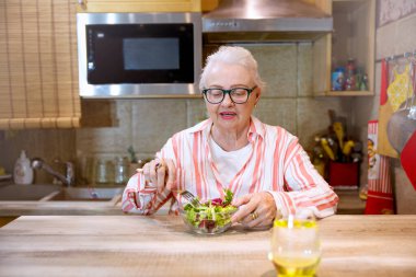 Sağlıklı atıştırmalıklar ve bir bardak su içen yaşlı kadın. Yaşlı kadın ahşap mutfak masasında taze salata yiyor. Boşluğu kopyala