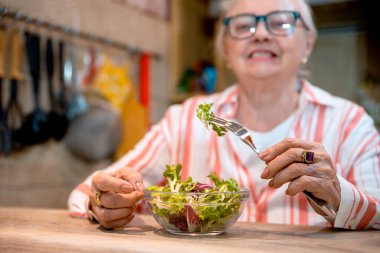 Sağlıklı atıştırmalıklar ve bir bardak su içen yaşlı kadın. Yaşlı kadın ahşap mutfak masasında taze salata yiyor. Boşluğu kopyala