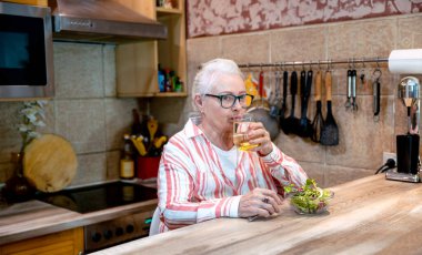 Sağlıklı atıştırmalıklar ve bir bardak su içen yaşlı kadın. Yaşlı kadın ahşap mutfak masasında taze salata yiyor. Boşluğu kopyala