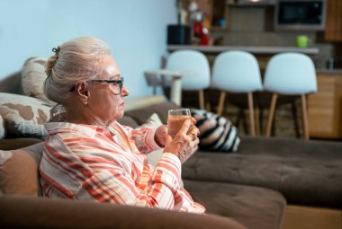 Kafkas kıdemli bayan dinlenirken bir bardak taze suyla kanepede oturuyor. Yaşlı beyaz saçlı büyükanne suyla ferahlatıyor.