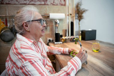 Sağlıklı atıştırmalıklar ve bir bardak su içen yaşlı kadın. Yaşlı kadın ahşap mutfak masasında taze salata yiyor. Boşluğu kopyala