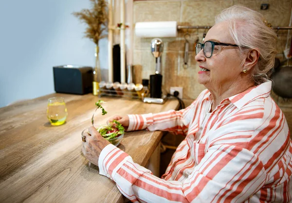 Sağlıklı atıştırmalıklar ve bir bardak su içen yaşlı kadın. Yaşlı kadın ahşap mutfak masasında taze salata yiyor. Boşluğu kopyala
