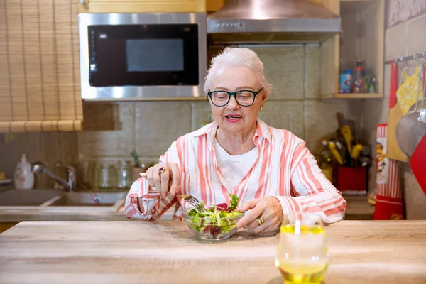 Sağlıklı atıştırmalıklar ve bir bardak su içen yaşlı kadın. Yaşlı kadın ahşap mutfak masasında taze salata yiyor. Boşluğu kopyala